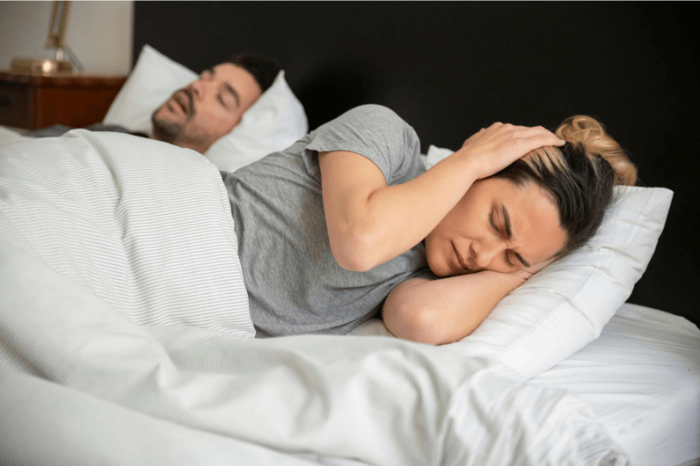 A couple in bed with one covering her ears, showing the impact of snoring